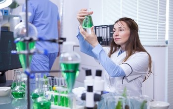 Mujer observando una sustancia de color verde en un matraz sentada frente a una mesa con materiales de laboratorio