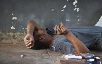 Hombre adicto a la drogas acostado en el suelo, su brazo izquierdo sobre el rostro con una jeringa en su mano y al lado celular y drogas
