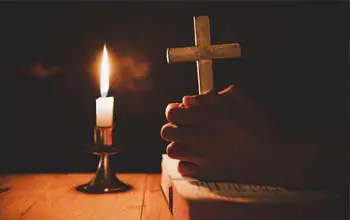 unas manos en posición de oración sosteniendo una cruz y una vela encendida