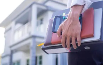 Mano de un freelance sosteniendo una calculadora, un libro y una carpeta de cuentas, en el fondo se ve una casa desenfocada.