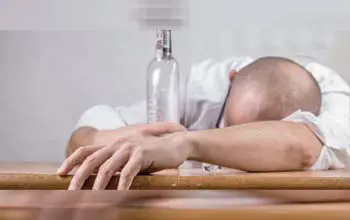 Hombre en posición de descanso sobre mesa de madera con botella de alcohol vacía en fondo gris
