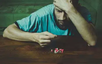 Hombre sentado frente a un grupo de pastillas sobre una mesa de madera con una mano en su frente y la otra tomando una pastilla