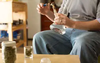 Hombre sentado encendiendo pipa con yesquero sentado frente a una mesa con dos frascos de marihuana en una sala