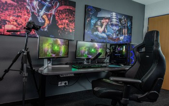 Se puede observar el set de un streamer donde esta el escritorio, una computadora con varios monitores, silla gamer, la camara y unos cuadros decorativos