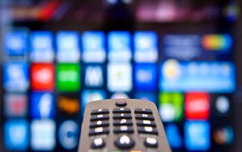 Un control remoto sostenido hacia una tv donde se ven, de forma borrosa, los logos de varias plataformas de streaming