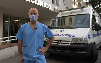 Enfermero vestido de azul con tapaboca parado afuera de centro clínico y al fondo una ambulancia