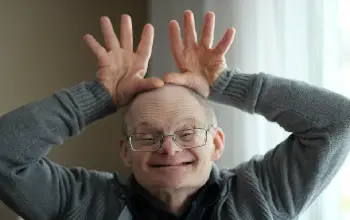 Hombre de tercera edad con síndrome de down sonriente con ambas manos abiertas sobre su cabeza parado en una sala