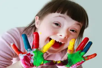 Niña con síndrome de down sonriendo mostrando las palmas de sus manos pintadas de varios colores en un fondo gris