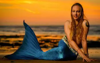 Mujer con cola de sirena de color azul sonriendo cabello largo sentada sobre la arena en la playa en un atardecer