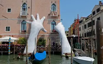 Escultura de dos manos gigantes de color blanco saliendo del agua tocando un edificio en una ciudad