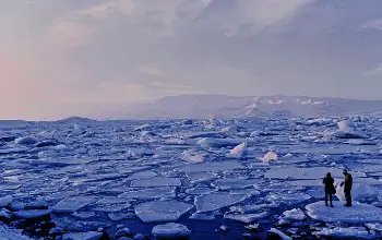 Dos personas paradas sobre un pedazo de hielo en el mar congelado con neblina y montañas al fondo
