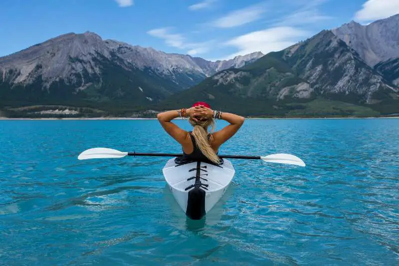 Mujer con sus manos sobre la cabeza sentada de espalda dentro de un kayak blanco y negro en el mar observando unas montañas frente a ella