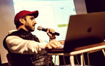 Hombre sentado hablando por un micrófono haciendo una presentación con una laptop y un proyector