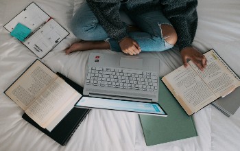 Persona sentada sobre una cama utilizando una laptop, libros y apuntes como fuente de información