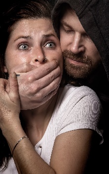 Hombre reflejando maldad en su rostro agarrando a una mujer por detrás y tapándole la boca en un fondo oscuro