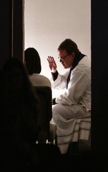 Sacerdote vestido con una Alba blanca sentado con la mano derecha levantada frente a una mujer confesándose sentada de espalda