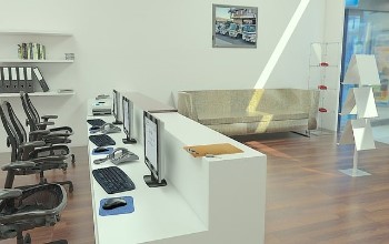 Puestos de trabajo en la recepción de una empresa de piso de madera y paredes blancas compuesta de computadoras, escritorio, sillas, sofá, etc