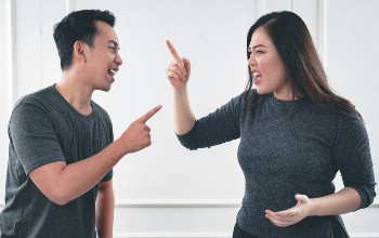 Hombre y mujer discutiendo señalándose mutuamente con el dedo indice en un fondo color blanco