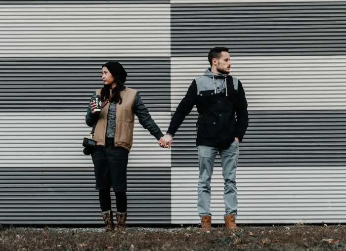 Hombre y mujer parados sobre un césped tomados de la mano, indiferentes sobre un fondo de zinc gris