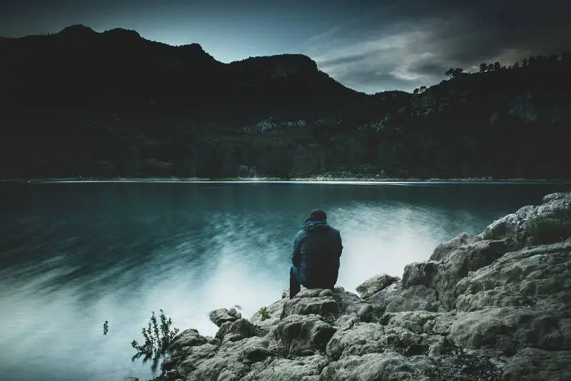 Hombre sentado sobre una roca enorme frente a un lago con montañas y arboles con el cielo nublado