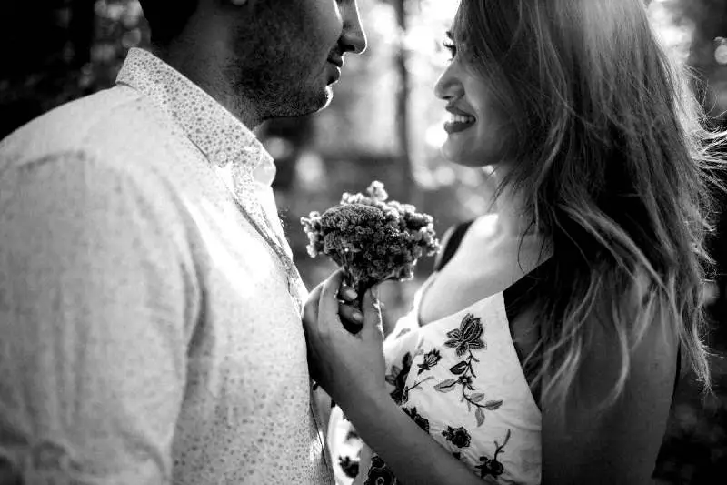 Foto en blanco y negro una mujer sosteniendo un ramito de flores parada mirando a los ojos a un hombre en un fondo borroso