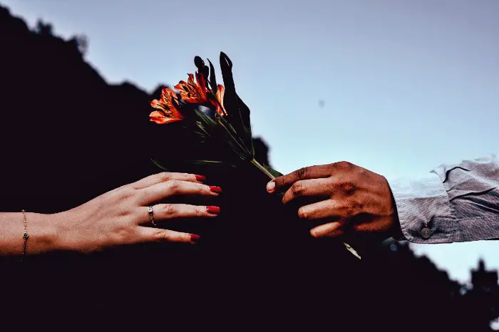 Imagen con la mano de un hombre con un ramo de flores entregándolo a una mano de mujer en un fondo borroso de atardecer
