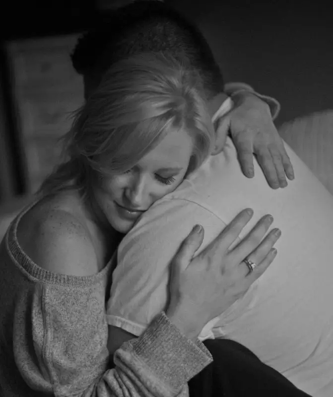 Foto en blanco y negro de mujer sentada abrazando a un hombre en un fondo borroso